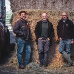 Dhruv Baker, Tom Whitaker and Andrew Owens stand in front of stacked bales of hay, with farming machinery visible to the left side of the image.